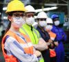 Fabrikarbeiterin mit Helm und Hygienemaske und Männer stehen in einer Fertigungsanlage Schlange.