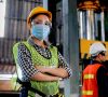 Arbeiterin trägt Mundschutz, eine Warnweste, einen Helm und eine Schutzbrille.