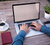 Eine Person arbeitet tippt auf die Tastatur eines Laptops. Neben dem Laptop stehen ein Kaffee, ein Glas und ein rotes Mäppchen.