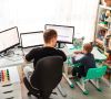 Ein Mann arbeitet im Kinderzimmer seines Sohnes, er sitzt an einem Schreibtisch mit drei Bildschirmen, sein Sohn sitz daneben an einem kleineren Schreibtisch.