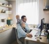 Ein Mann arbeitet zu Hause an seinem Laptop und hält sein Baby.