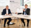 Holger Hanselka (links), Präsident KIT, und Heinz-Jürgen Prokop, CEO Trumpf Werkzeugmaschinen, sitzen an einem Tisch. Prokop hält den Kooperationsvertrag in die Höhe.